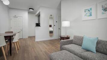A modern living room with a grey sofa, a dining table with chairs, a mirror, and a lamp.