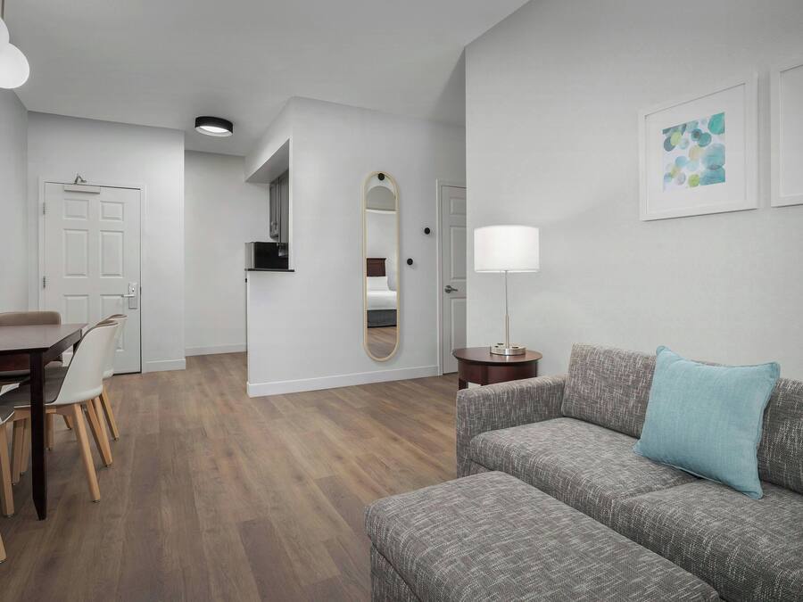 A modern living room with a grey sofa, a dining table with chairs, a mirror, and a lamp.