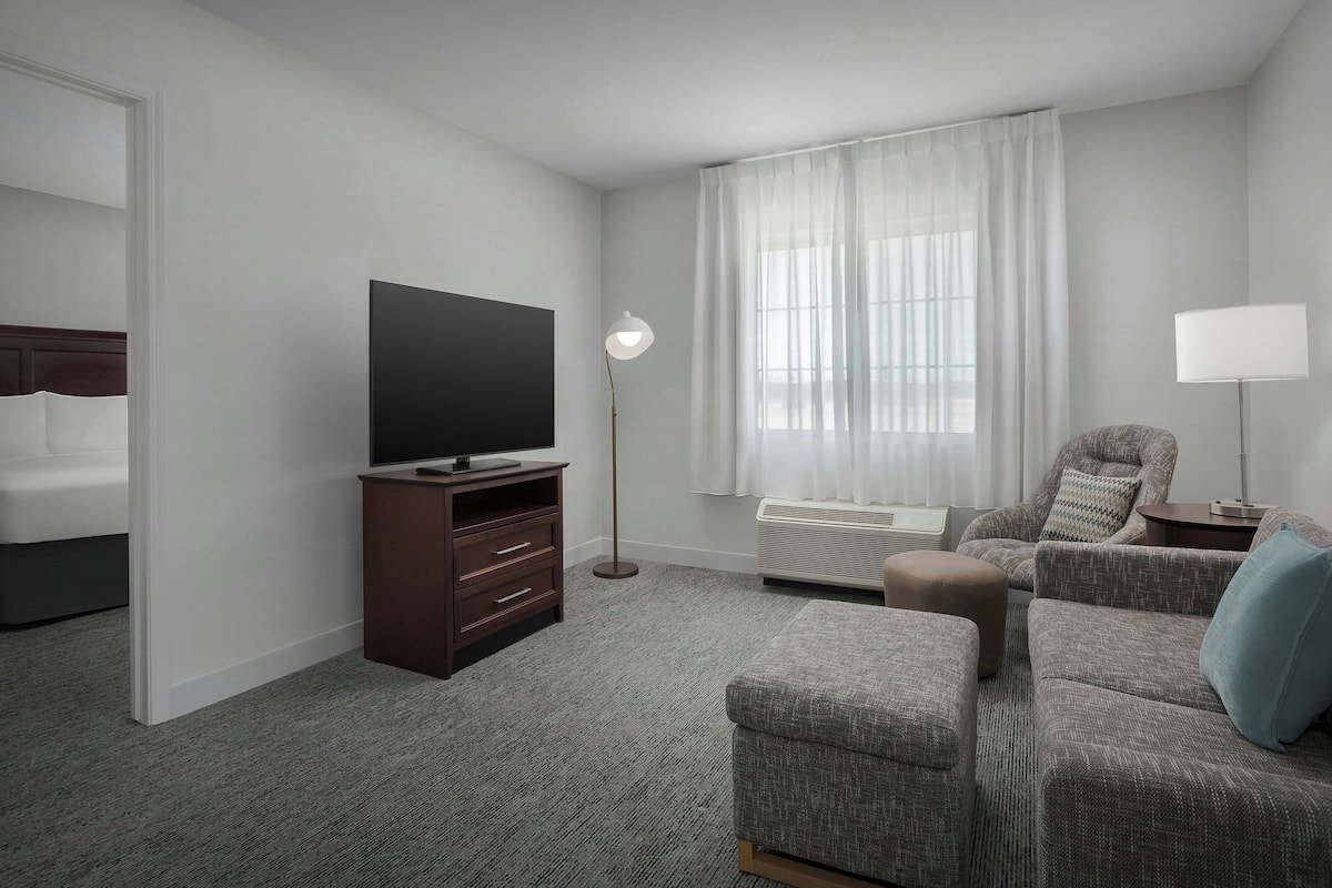 A hotel room with a flat-screen TV, a dark wooden dresser, a grey sofa, a patterned armchair, a floor lamp, and a large window with sheer curtains.