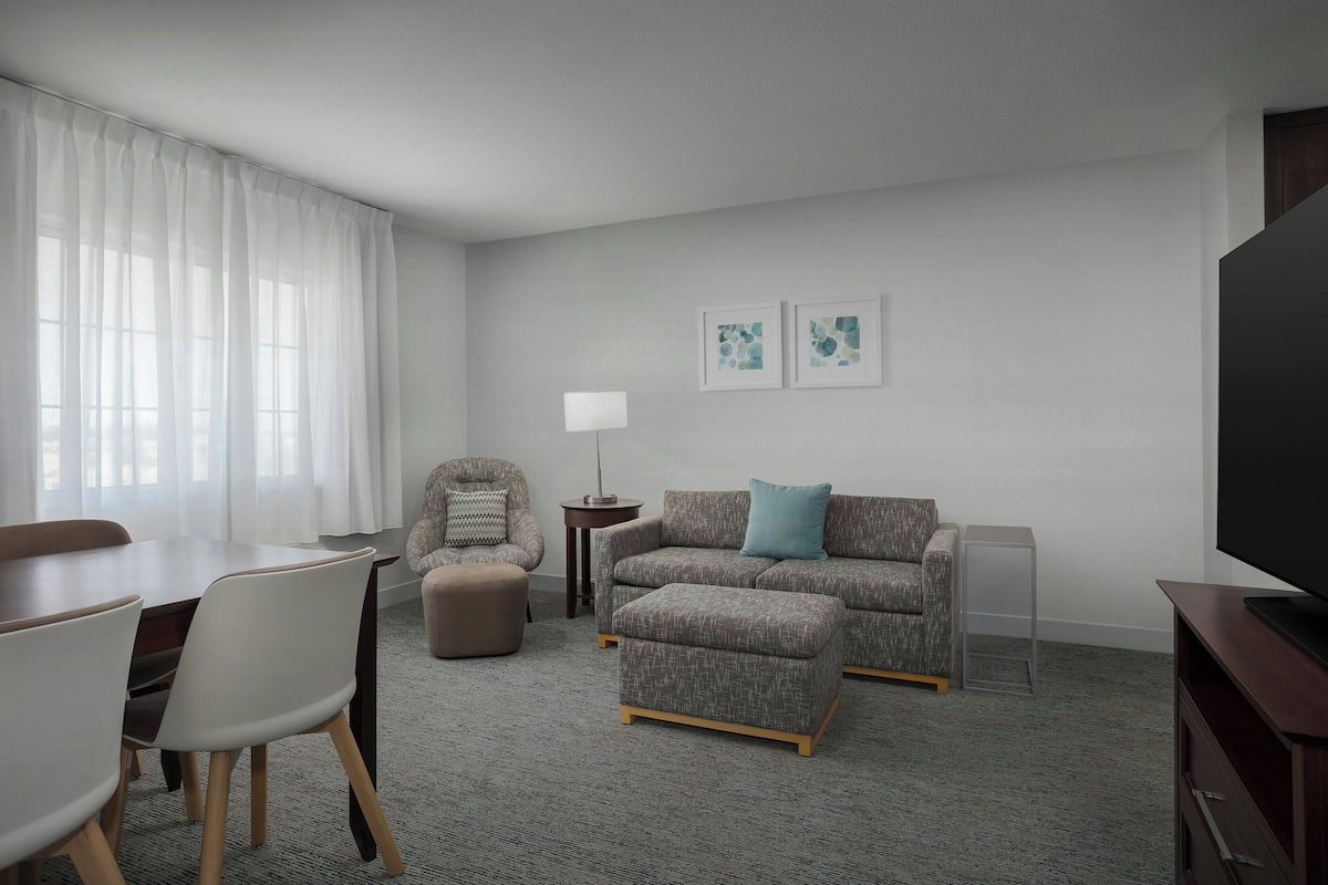 A modern living room with a dining area, grey carpet, a grey sofa with a blue pillow, a round table with chairs, a lamp, and two framed pictures on the wall.