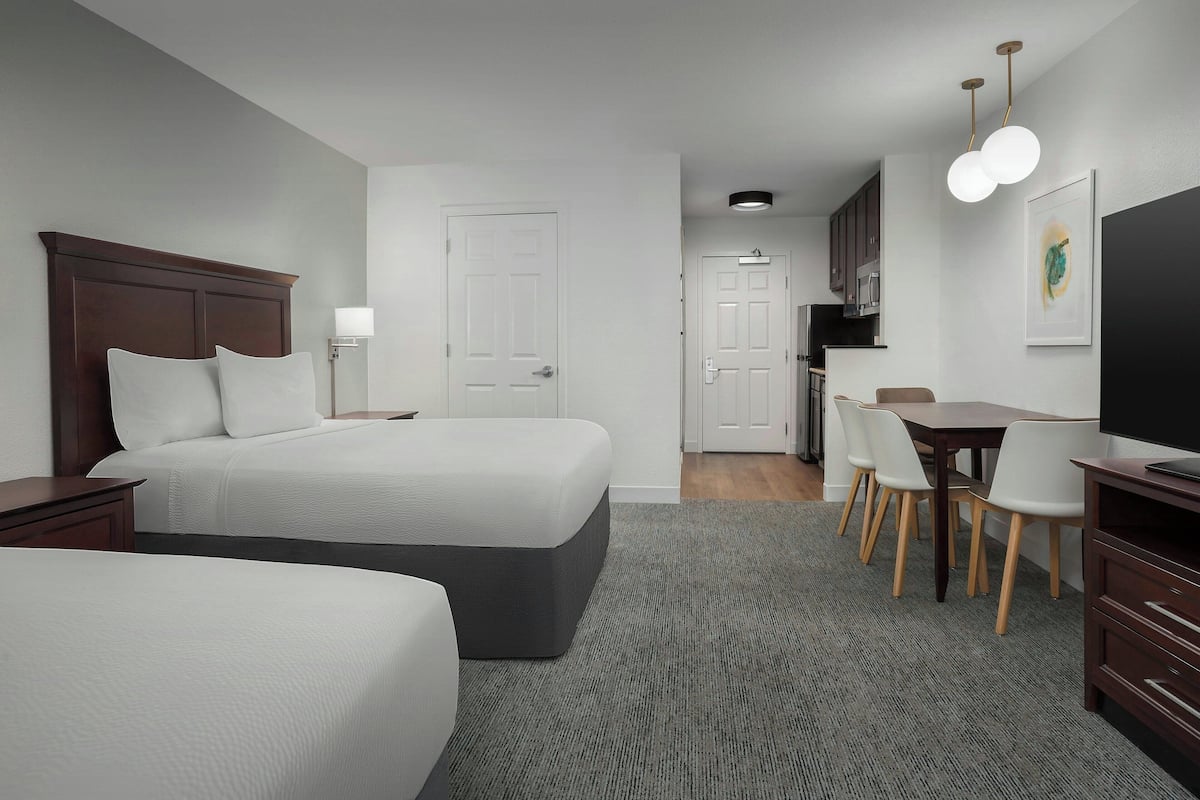 A hotel room with two beds, a desk, chairs, a television, and a kitchenette.