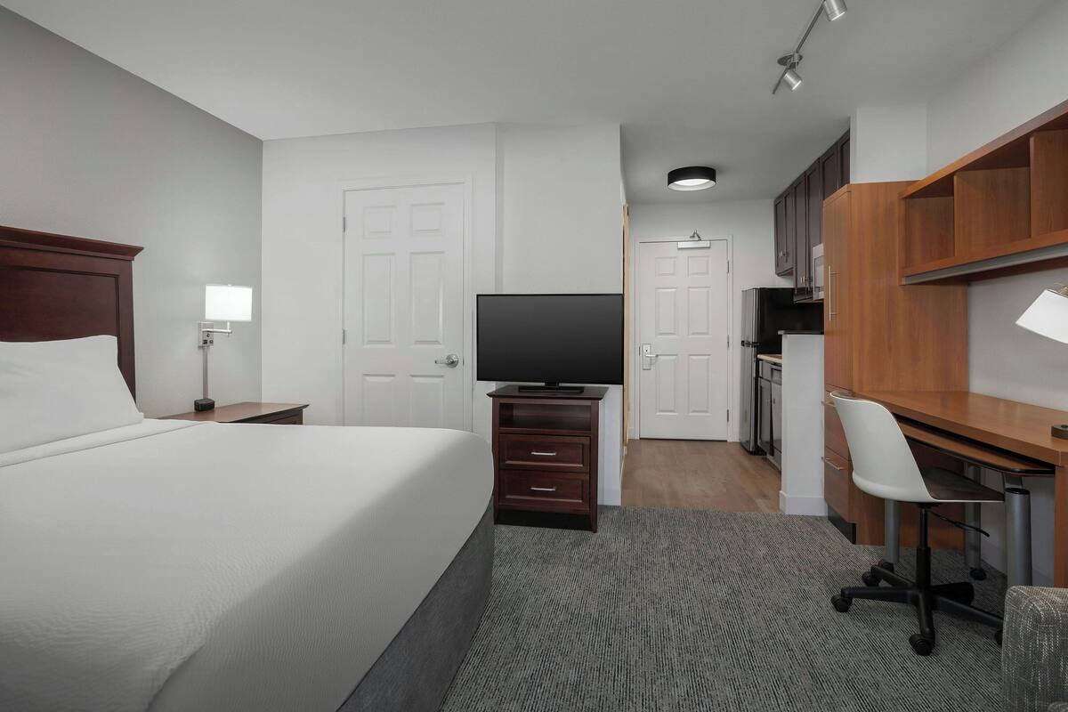 A hotel room with a bed, a TV, a desk with a chair, and a kitchenette.
