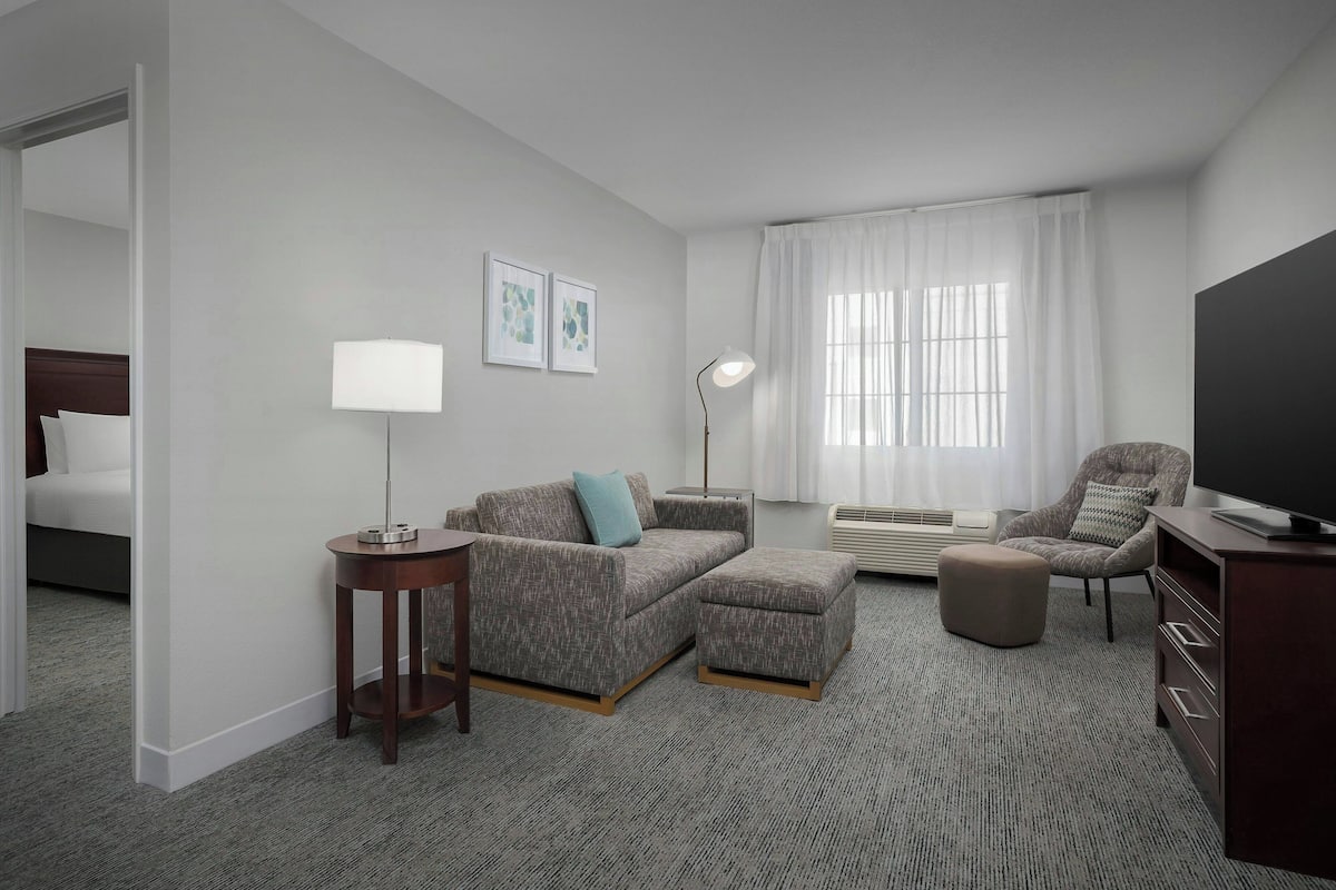 A hotel room with a grey sofa set, a small table, a chair, a TV, and a large window with curtains.