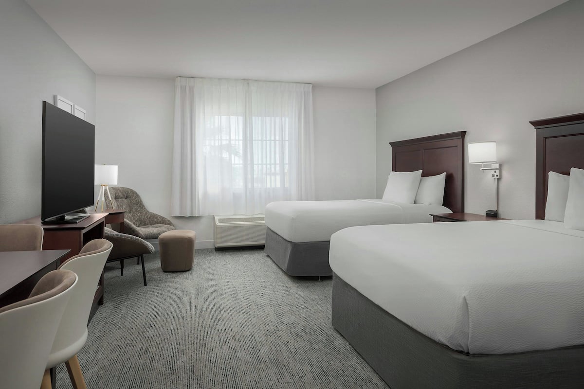 A hotel room with two beds, a desk with a TV, a chair, a sofa, and a large window with curtains.