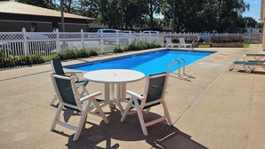 A swimming pool with lounge chairs and a table, surrounded by a white fence and greenery.