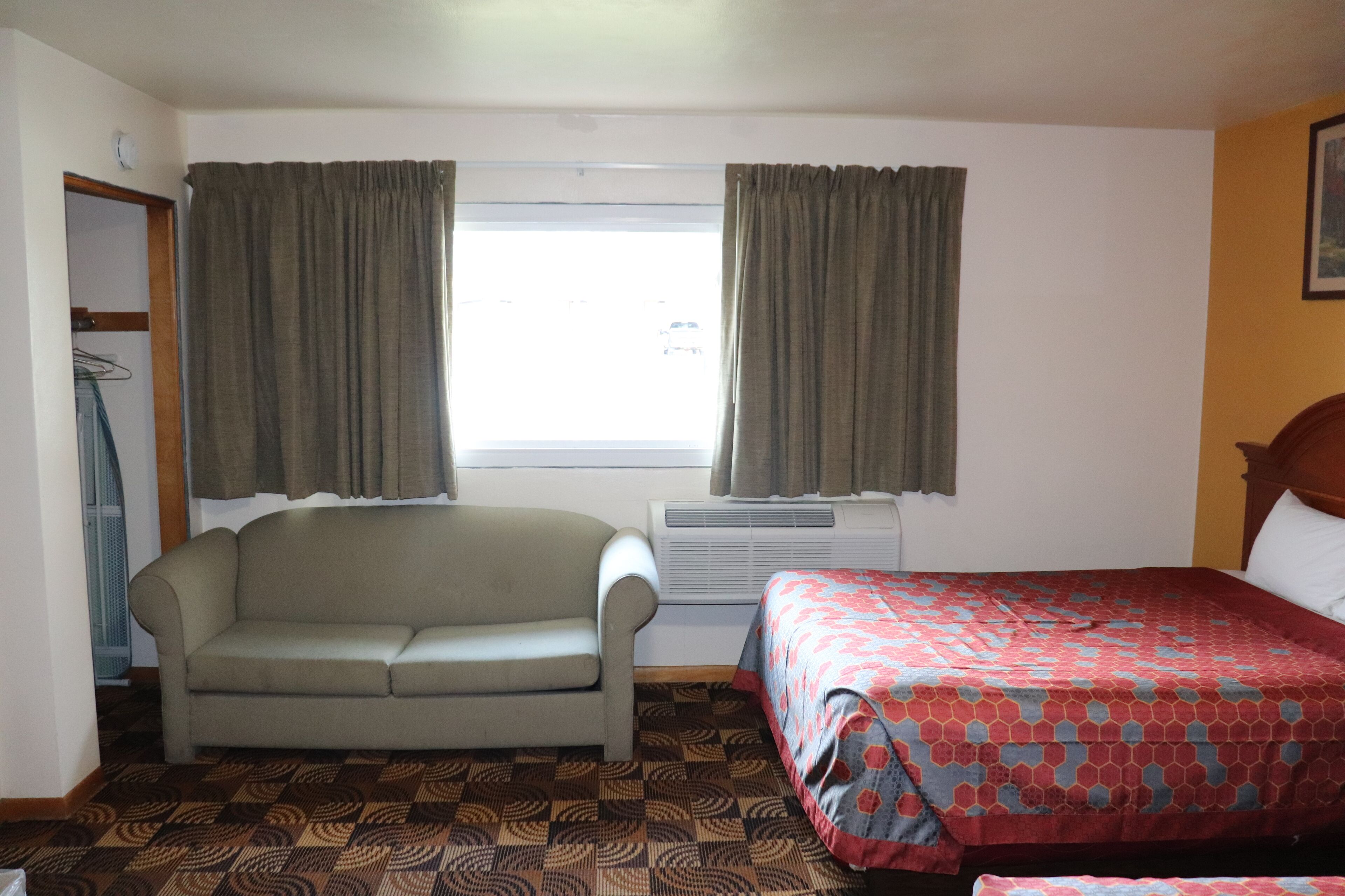 A hotel room with a sofa, a bed with a patterned bedspread, a window with curtains, and a radiator.