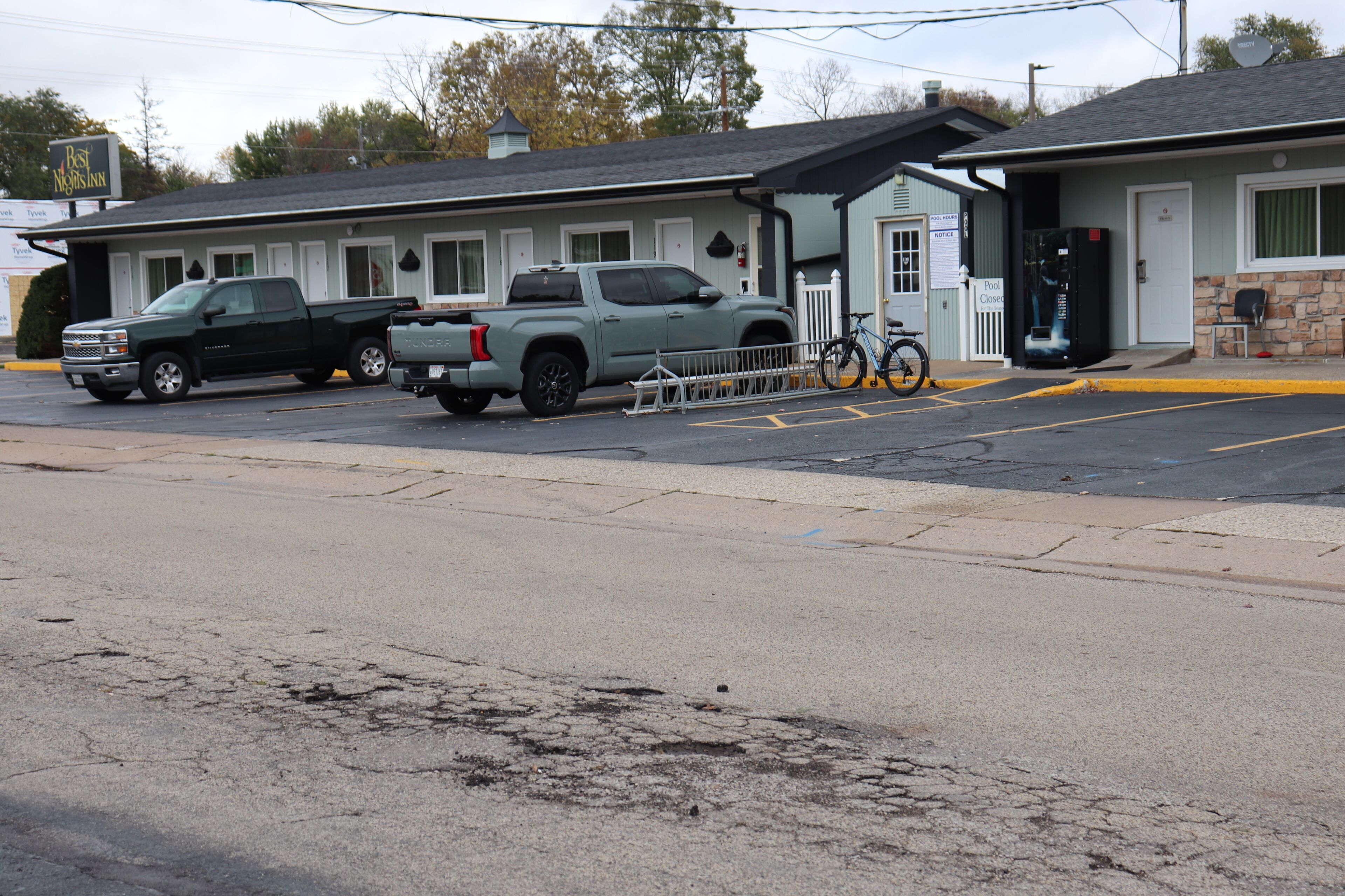 Un motel avec deux camions garés, un vélo et un panneau indiquant les heures d’ouverture de la piscine.