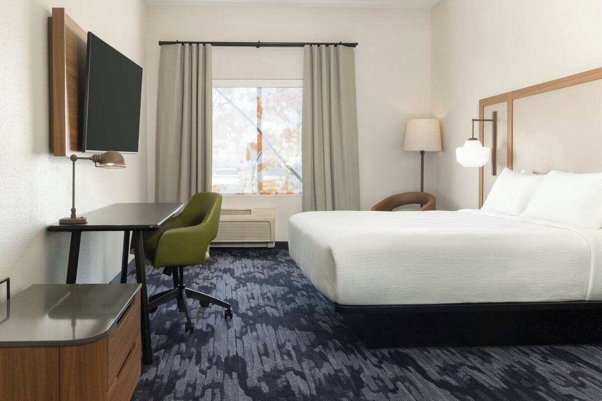 A hotel room with a bed, desk, chair, TV, lamp, and window with curtains.