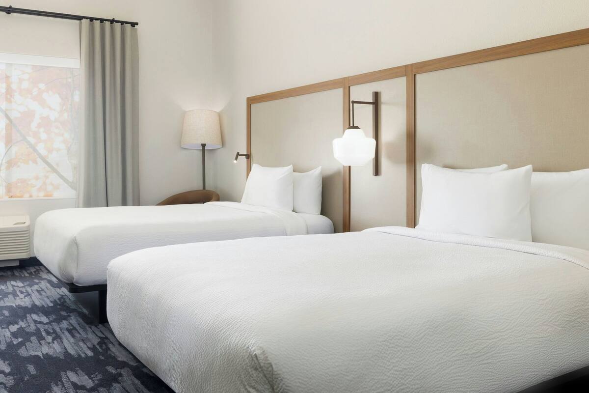 A hotel room with two beds, a window with curtains, a lamp, and a wall-mounted light fixture.
