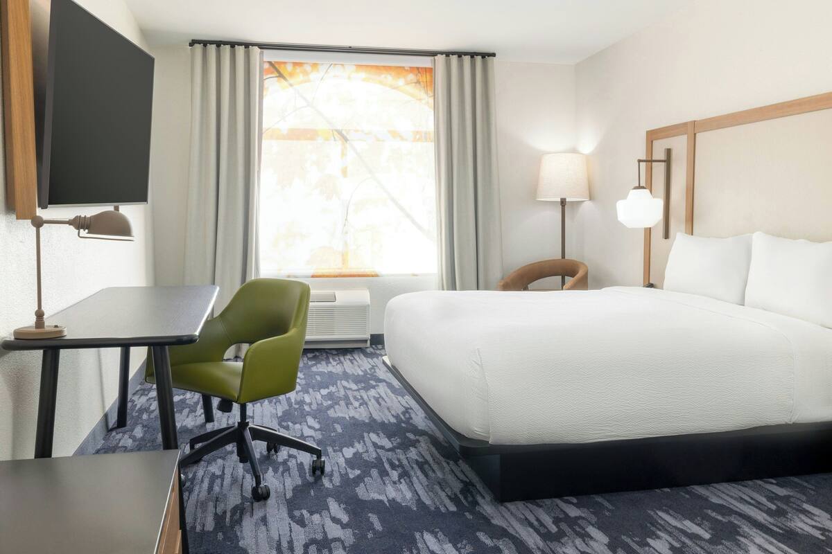A hotel room with a bed, a desk, a chair, a TV, a lamp, and a window with curtains.