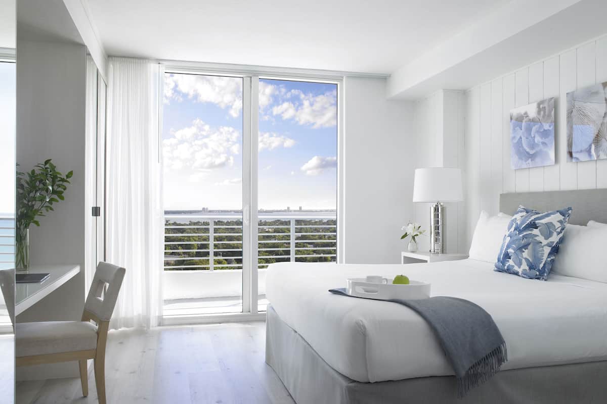 A modern hotel room with a large bed, a desk, and a view of the city skyline through floor-to-ceiling windows.