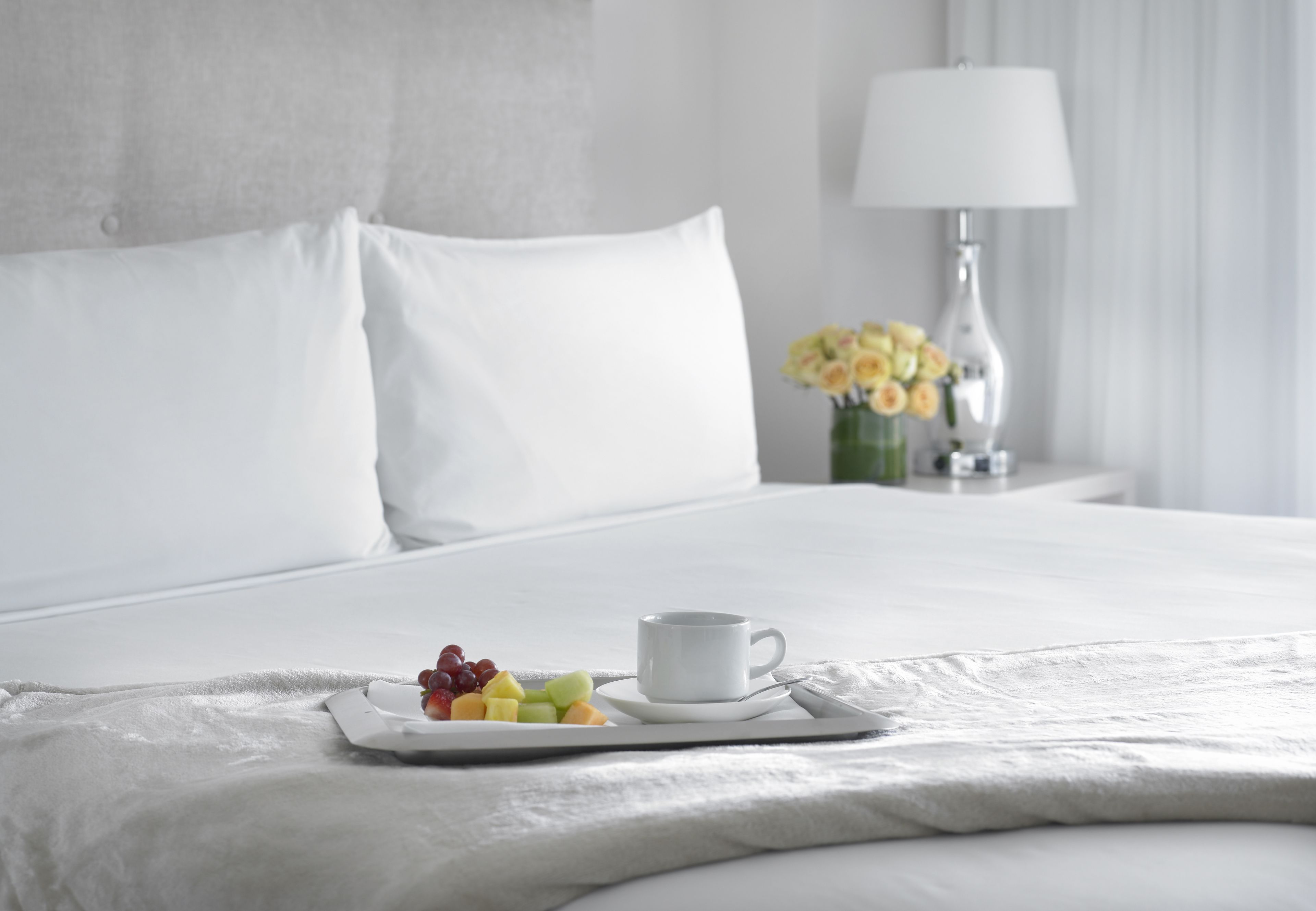 Uma cama bem arrumada com roupa de cama branca, uma bandeja com frutas e uma xícara de café, e um vaso com flores amarelas em uma mesa de cabeceira.