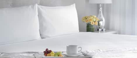 A neatly made bed with white bedding, a tray with fruit and a cup of coffee, and a vase with yellow flowers on a bedside table.