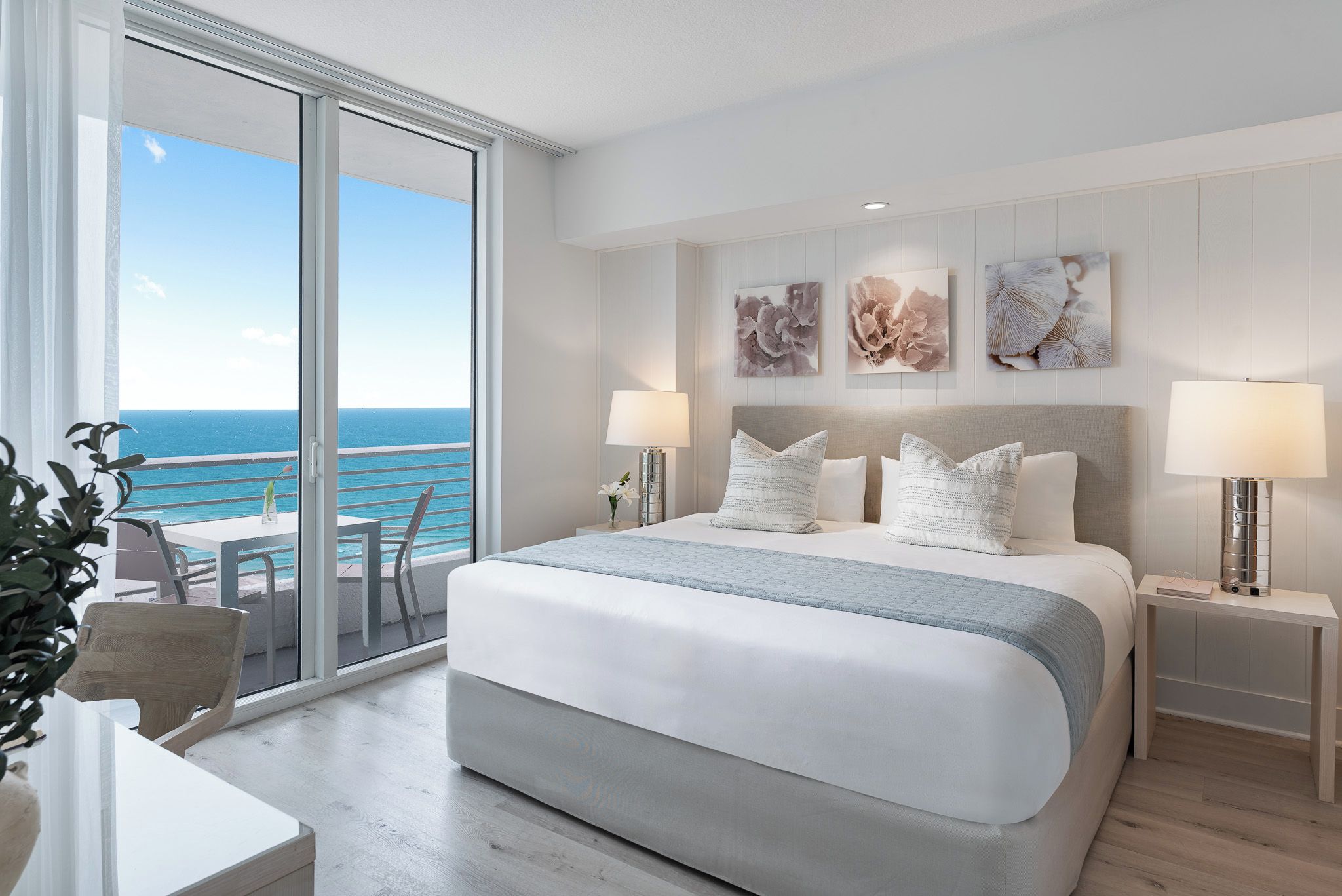 A hotel room with a large bed, two bedside lamps, a view of the ocean, and a balcony with a chair and table.