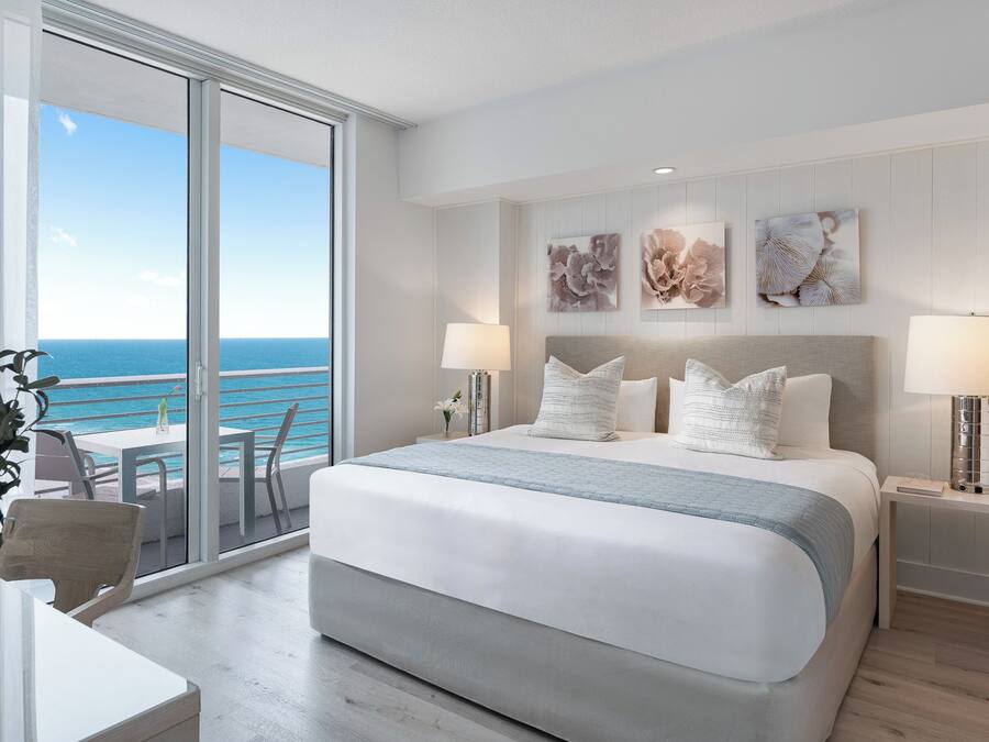 A hotel room with a large bed, two bedside lamps, a view of the ocean, and a balcony with a chair and table.