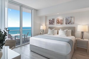 Habitación de hotel con una cama grande, dos lámparas de noche, vista al océano y un balcón con una silla y una mesa.