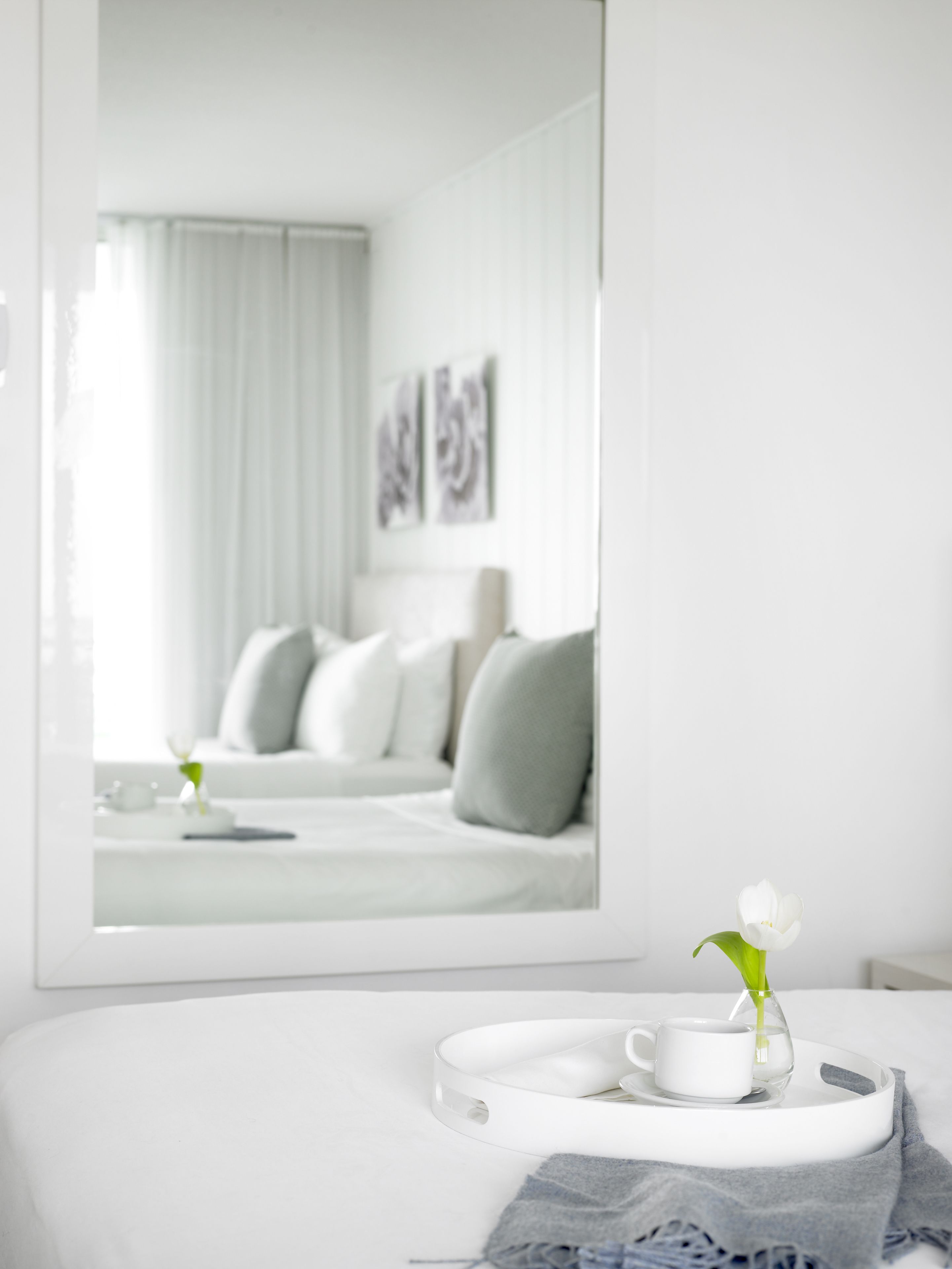 Camera d'albergo moderna e minimalista con un letto, un comodino con una tazza e un fiore, e un grande specchio che riflette l'ambiente.