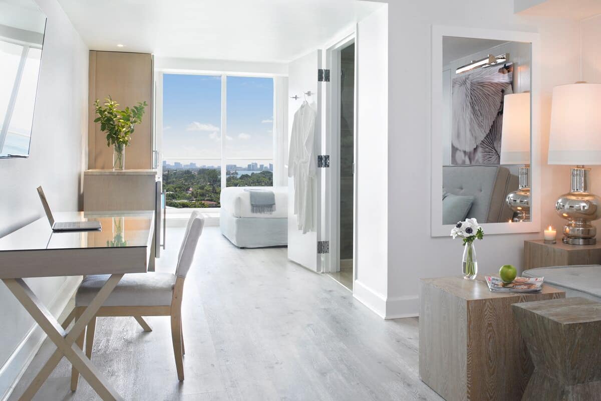 A modern living room with a sofa, a coffee table, a desk with a laptop, and a view of the city through large windows.