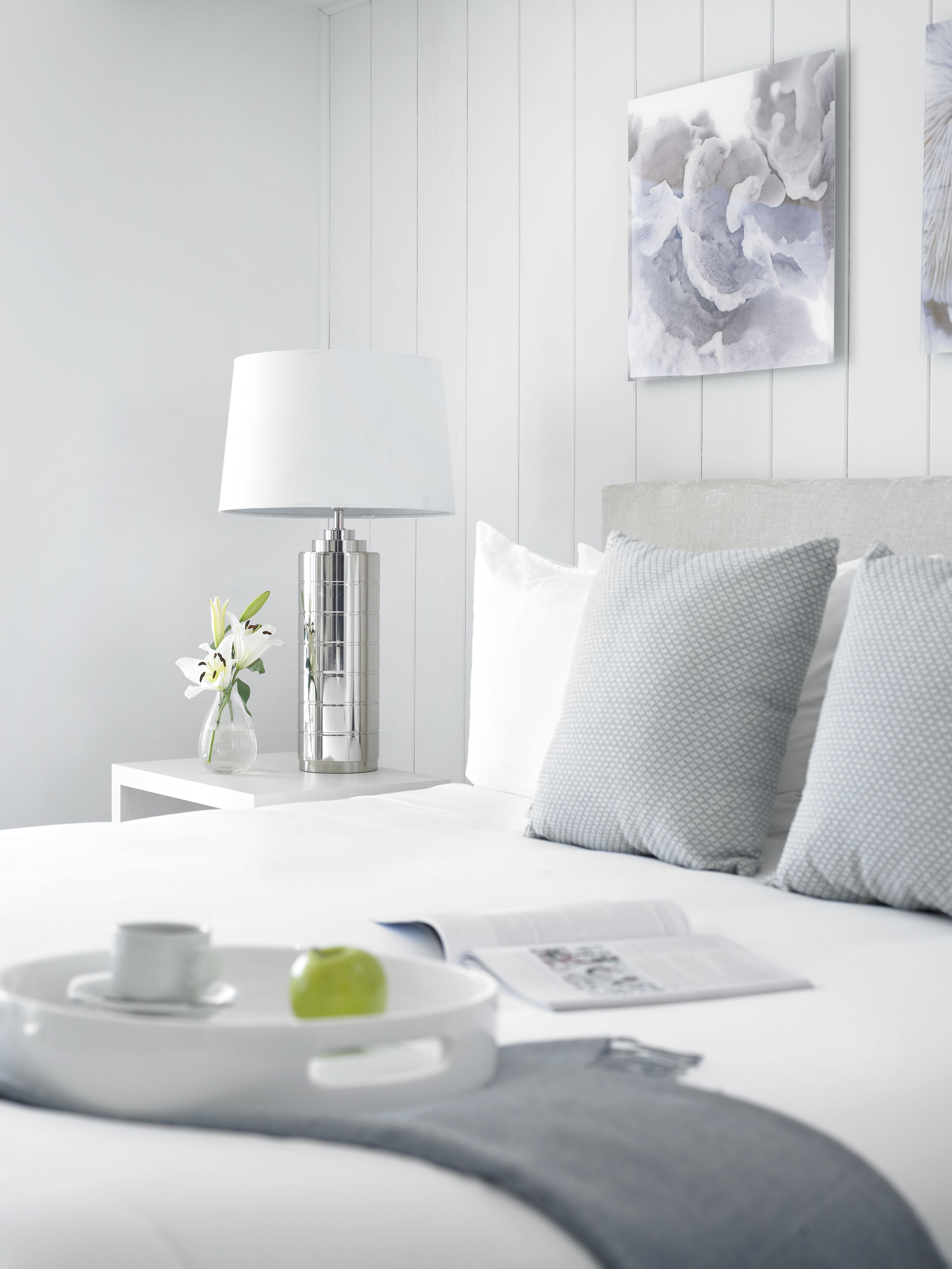 A modern bedroom with a white bed, a bedside table with a lamp, and a framed artwork on the wall.
