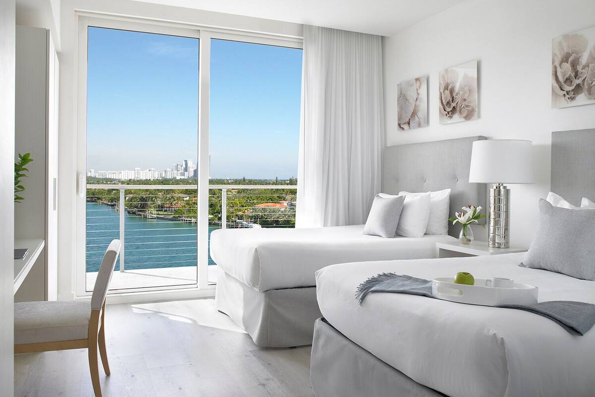 A modern hotel room with a large balcony, two beds, a desk, and a view of a city skyline and water.
