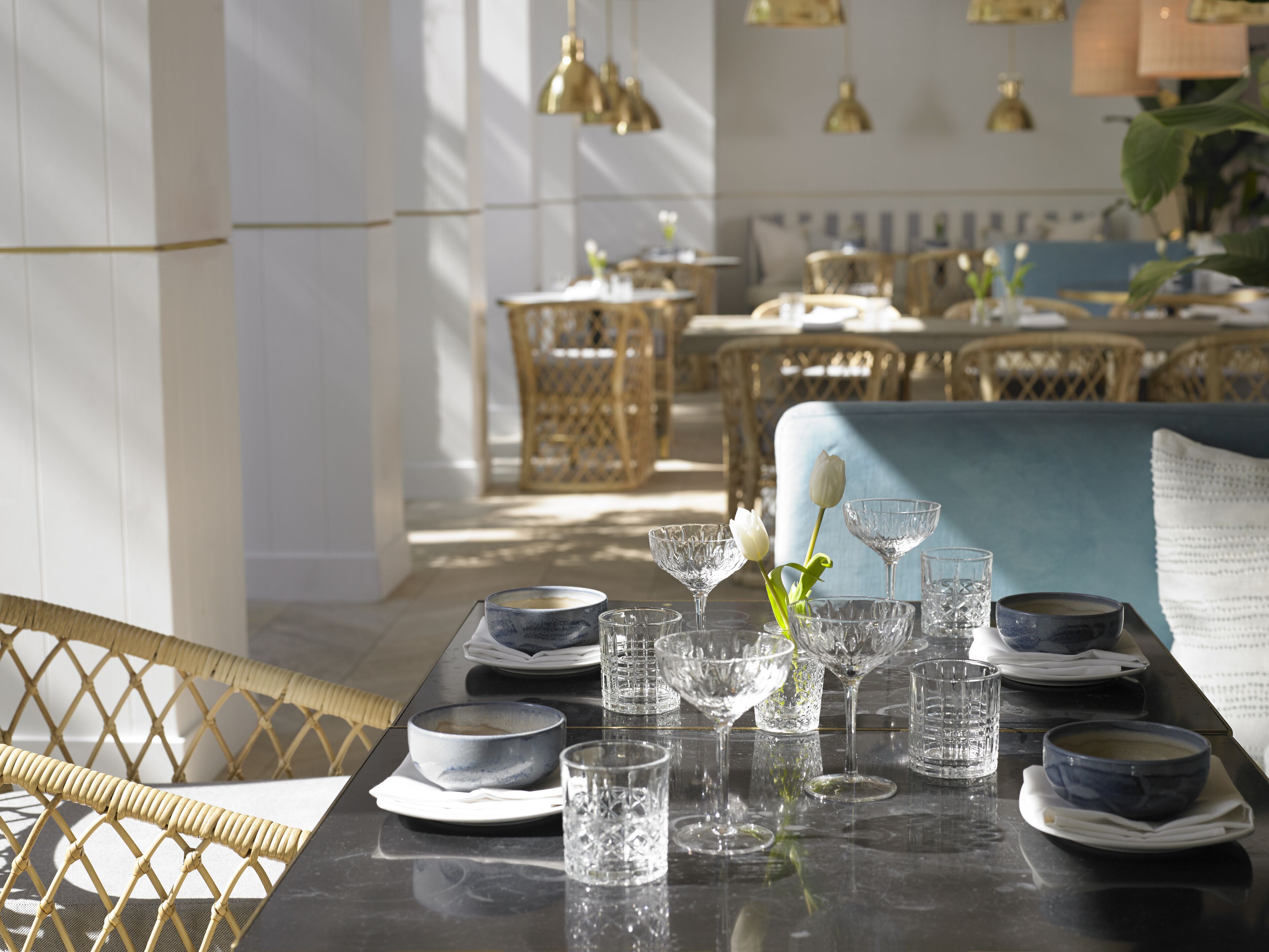 A dining area with a table set for four, featuring clear glassware and plates, a vase with a white flower, and wicker chairs.