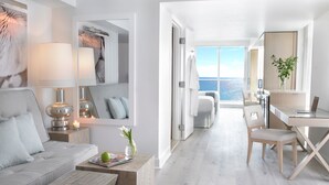 A modern, minimalist hotel room with a large window offering a sea view, a cozy seating area with a sofa and a wooden coffee table, and a small workspace with a laptop. - Grand Beach Hotel (Miami Beach)