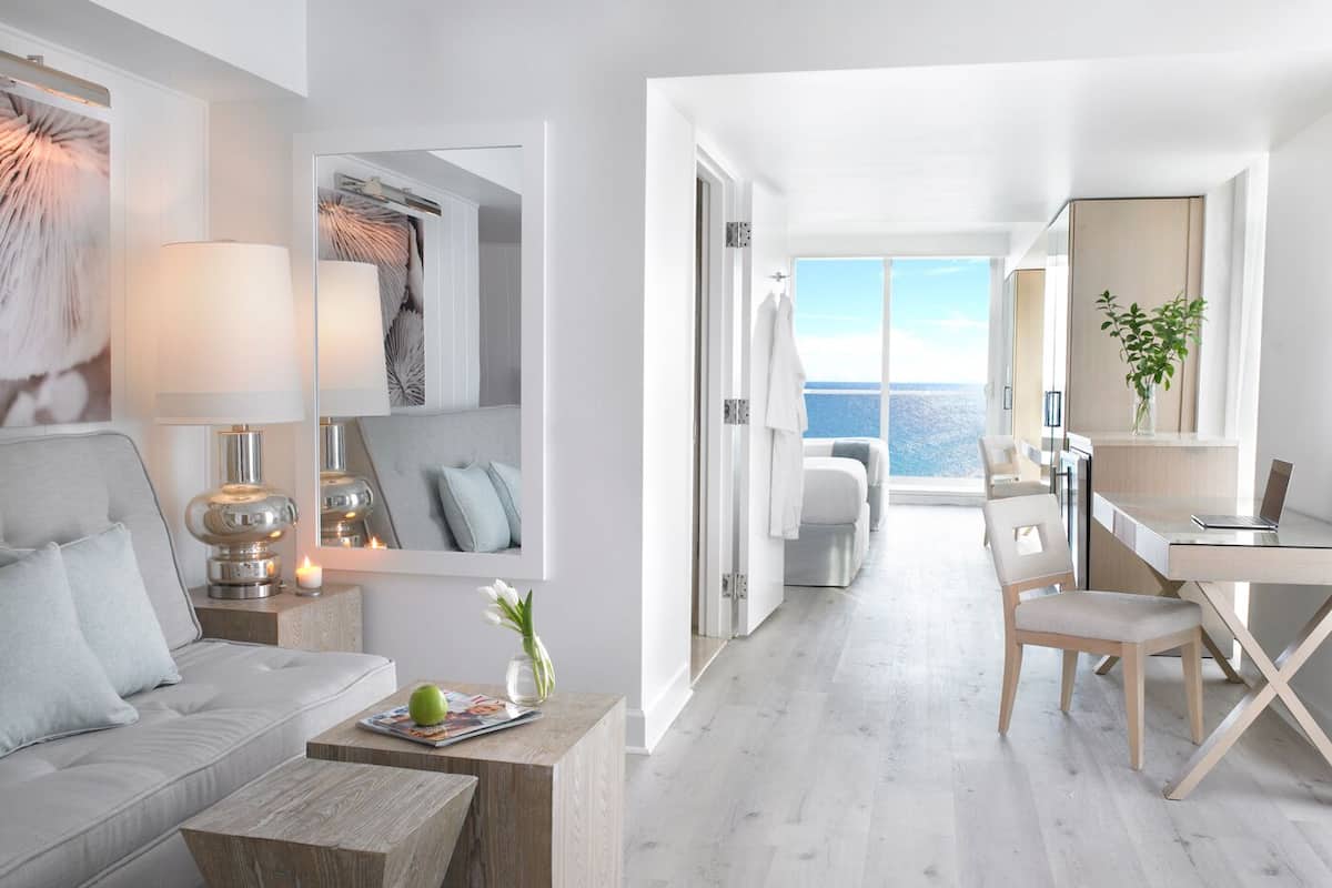 A modern, minimalist hotel room with a large window offering a sea view, a cozy seating area with a sofa and a wooden coffee table, and a small workspace with a laptop.