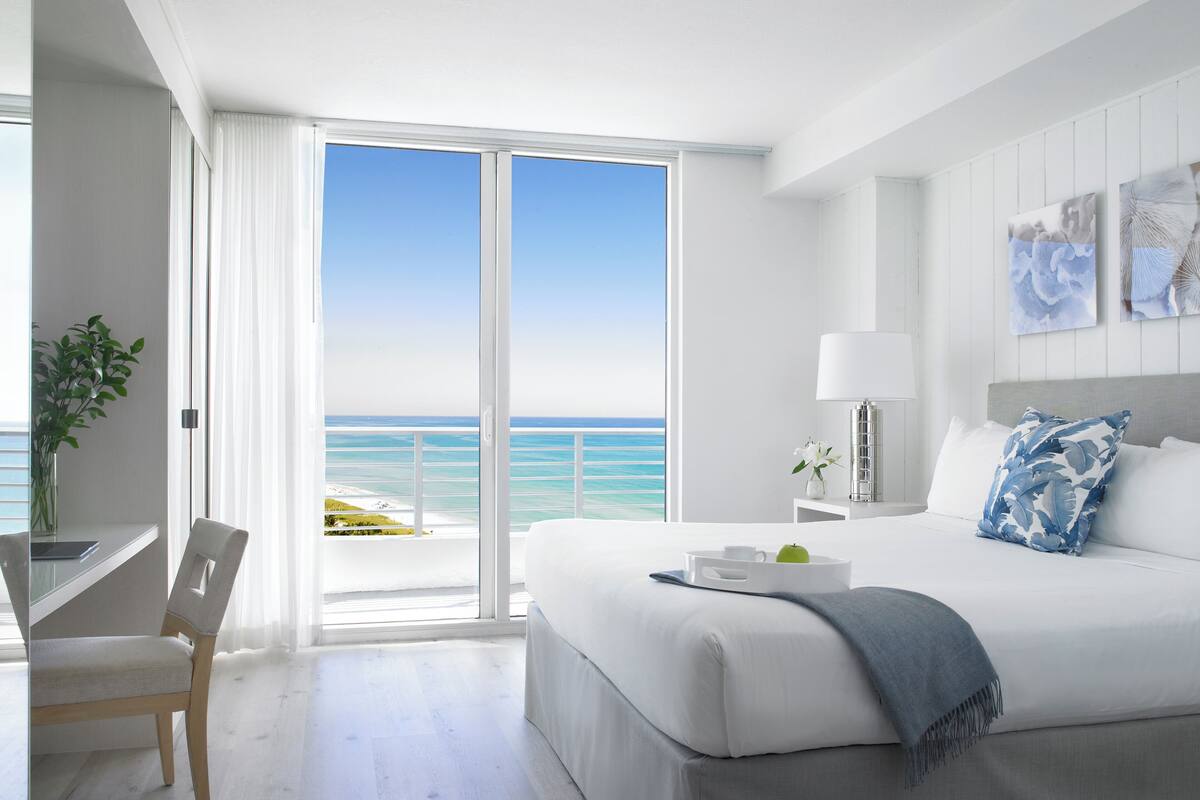A modern hotel room with a large bed, a desk, and a view of the ocean.