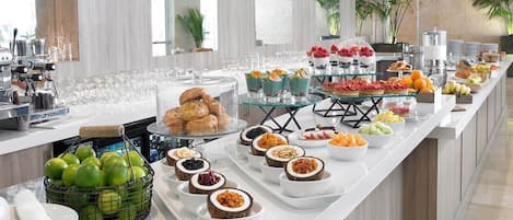 A buffet with various food items and beverages, including fruits, pastries, and a coffee station.