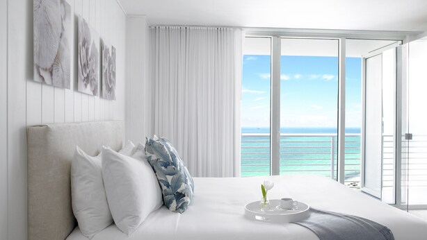 A hotel room with a large bed, white bedding, a view of the ocean, and a balcony with white curtains. - Grand Beach Hotel (Miami Beach)