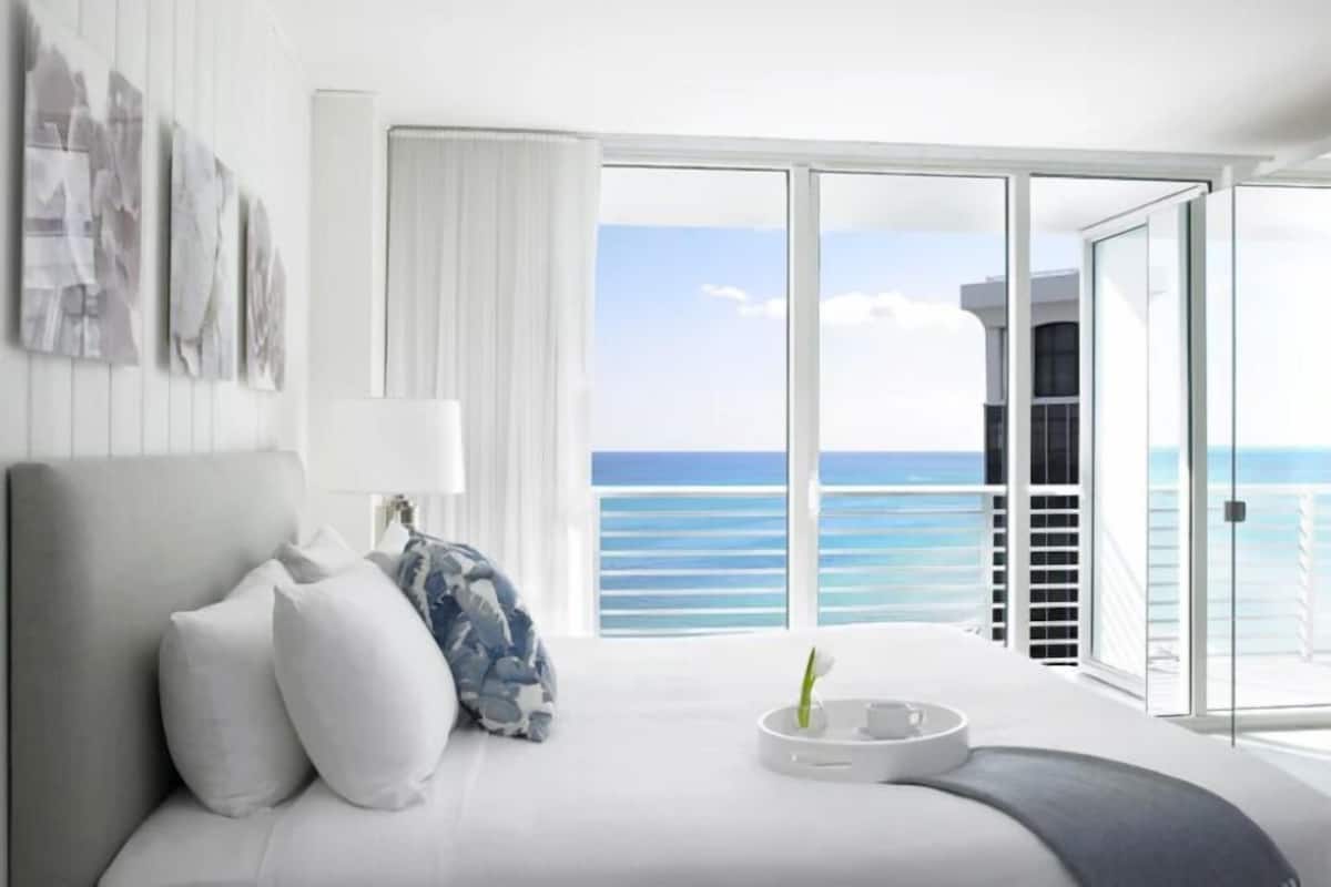 A hotel room with a bed, a bedside table with a vase, a large window with a view of the ocean, and a balcony with a railing.