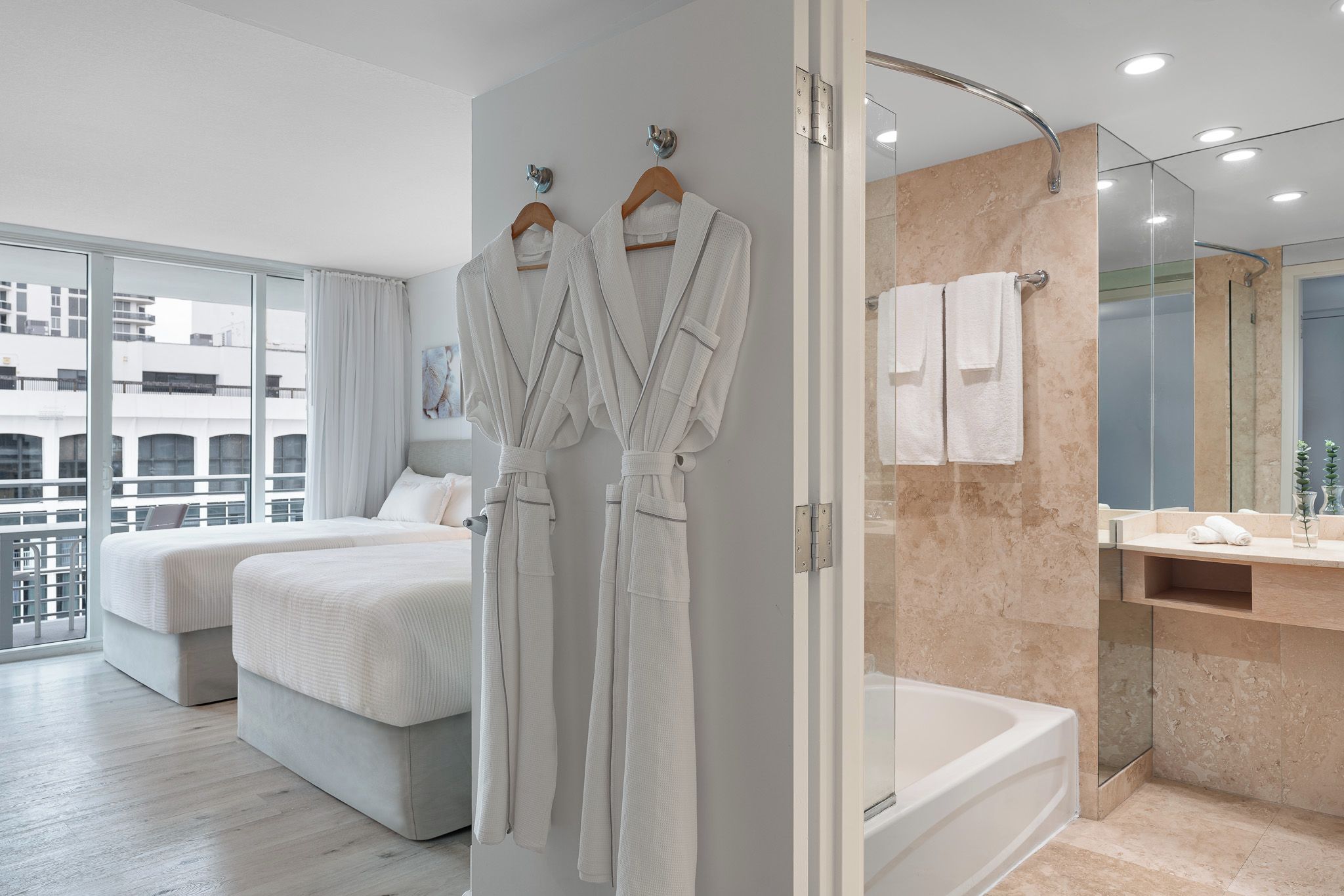 A modern hotel room with a large bed, a separate sitting area, a glass shower enclosure, and a view of the cityscape.
