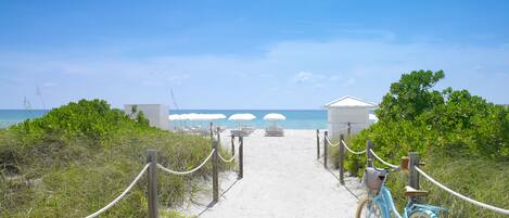 A sandy pathway leading to beach umbrellas and a small building, with a bicycle parked on the right.