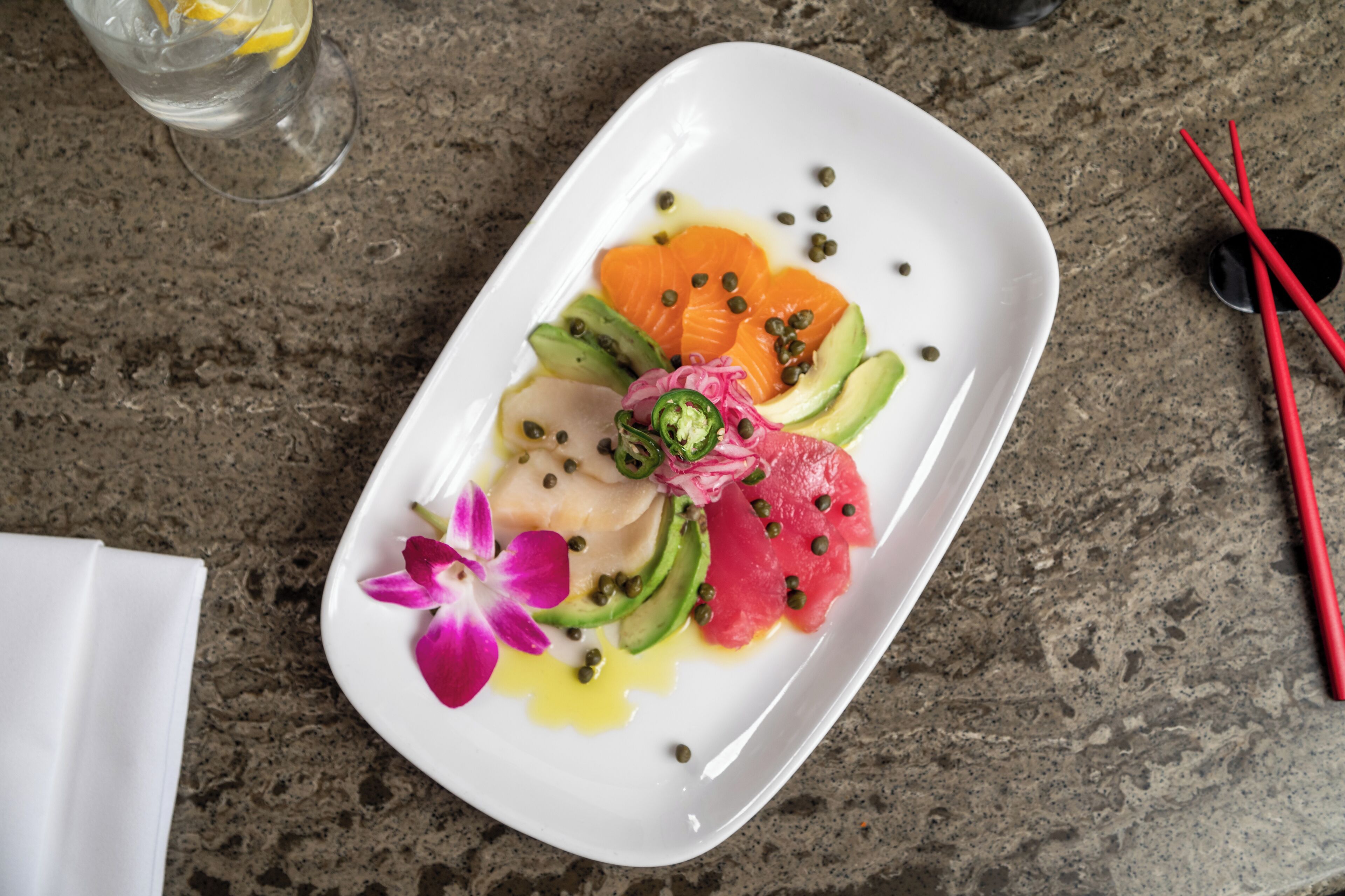 A white rectangular plate with various food items including fish, avocado, and a flower, placed on a granite countertop.