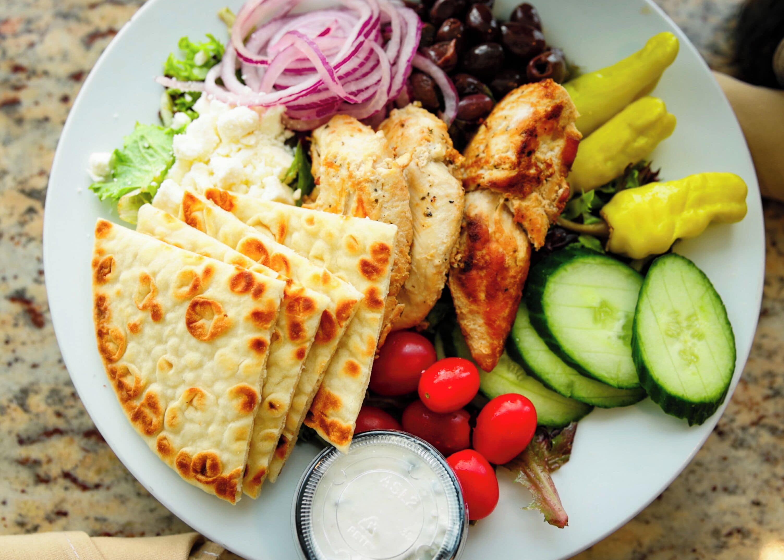 A plate with grilled chicken, pita bread, cucumbers, tomatoes, olives, and red onions.