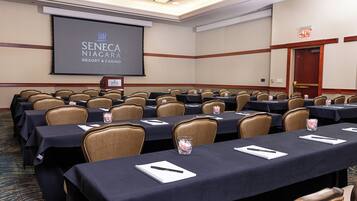 Een congrescentrum met rijen tafels en stoelen, een projectiescherm waarop het logo van Seneca Niagara Resort & Casino staat, en een deur met een vluchtwegbord.