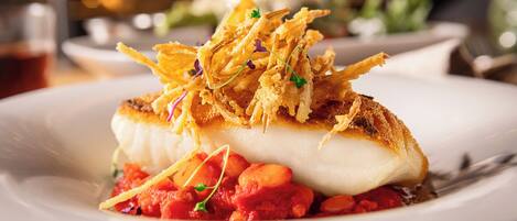 Un plato de comida con un filete de pescado blanco, adornado con tiras crujientes y hierbas, acompañado de salsa roja y frijoles.