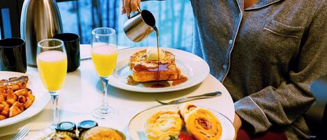 A breakfast table with various dishes, a coffee pot, and a person pouring syrup.