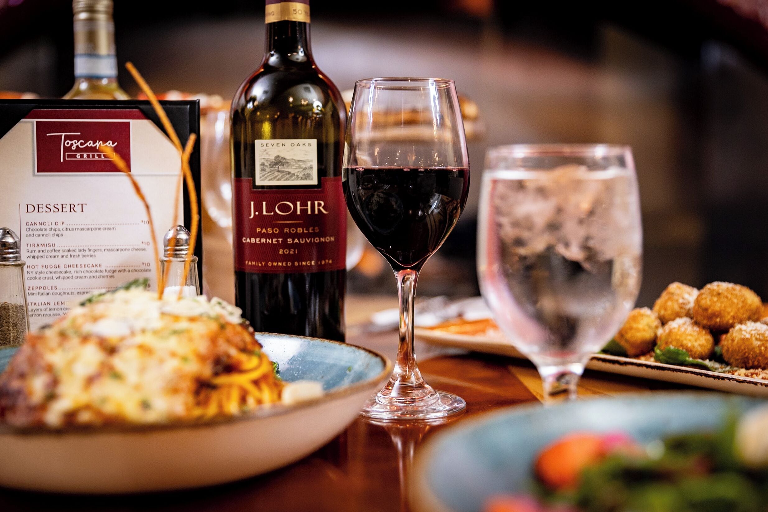 Una mesa de comedor con un menú, una botella de vino, una copa de vino y diversos platos.