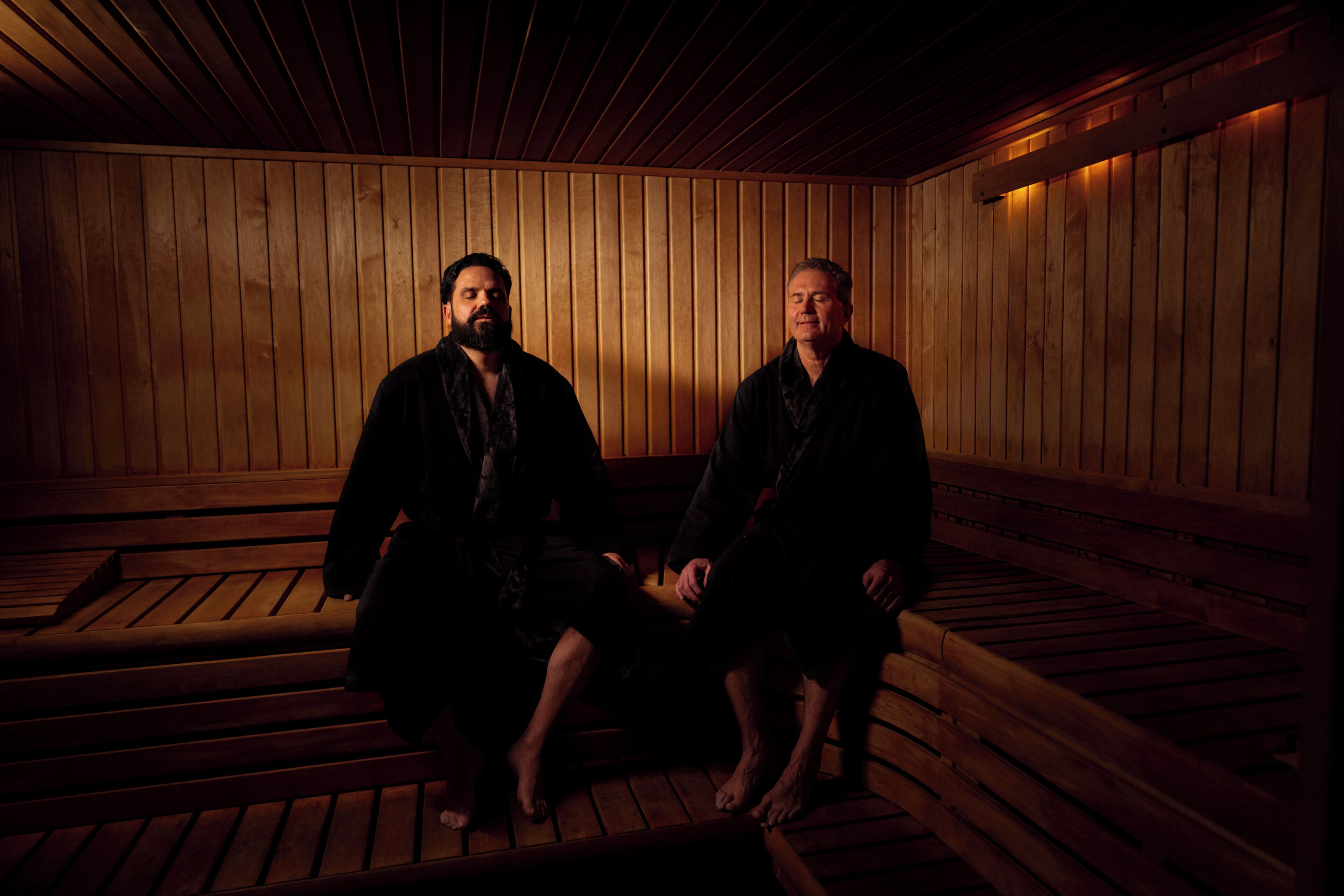 Dos personas sentadas en una sauna de madera con bancos y techo inclinado.