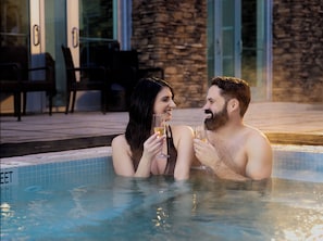 A couple in a hot tub with a stone wall and chairs in the background.