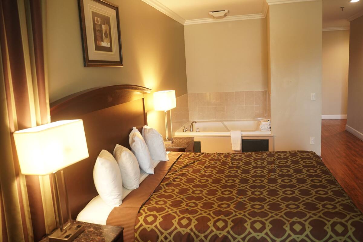 A hotel room with a large bed, a nightstand with a lamp, a bathroom with a bathtub, and a framed picture on the wall.
