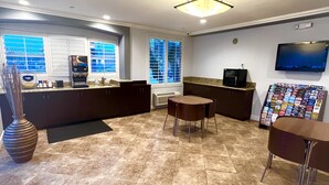 A modern kitchen with dark cabinetry, a central island, a flat-screen TV mounted on the wall, and a window with blinds. - Harbor Inn & Suites (Garden Grove)