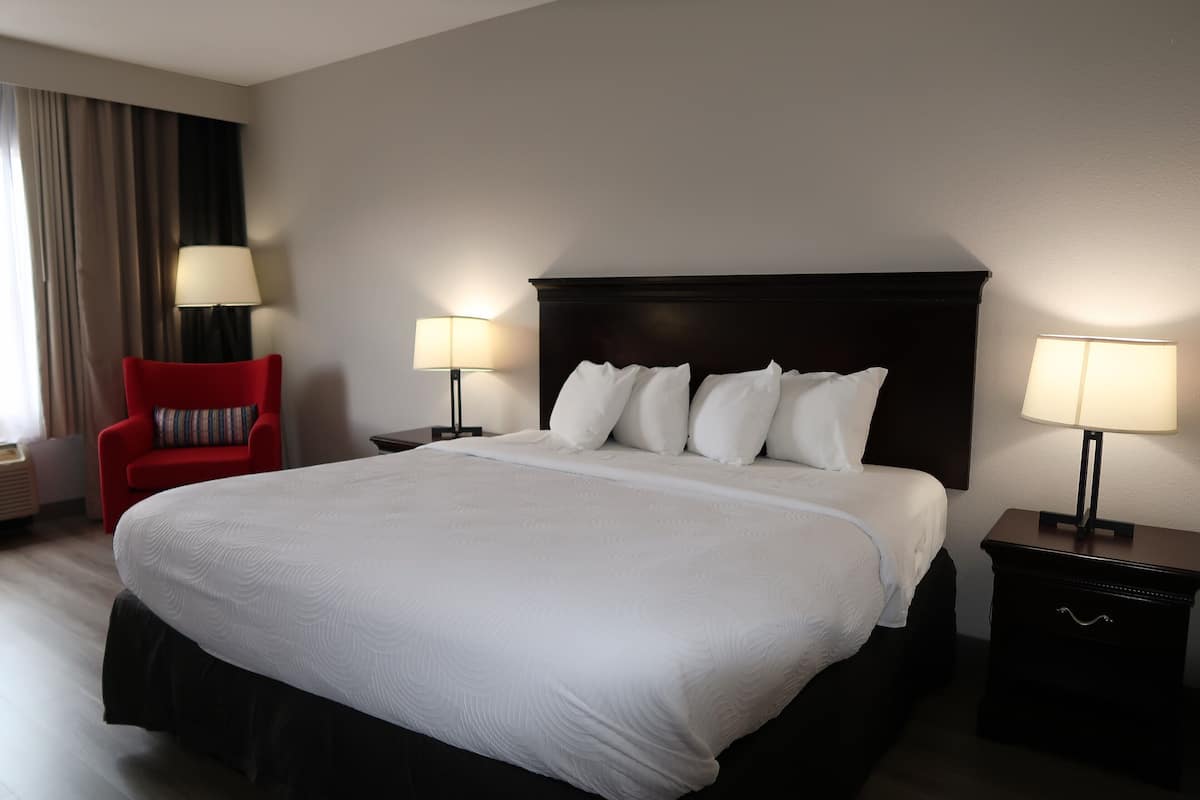 Habitación de hotel con una cama grande, dos mesitas de noche con lámparas, un sillón rojo y una ventana con cortinas.