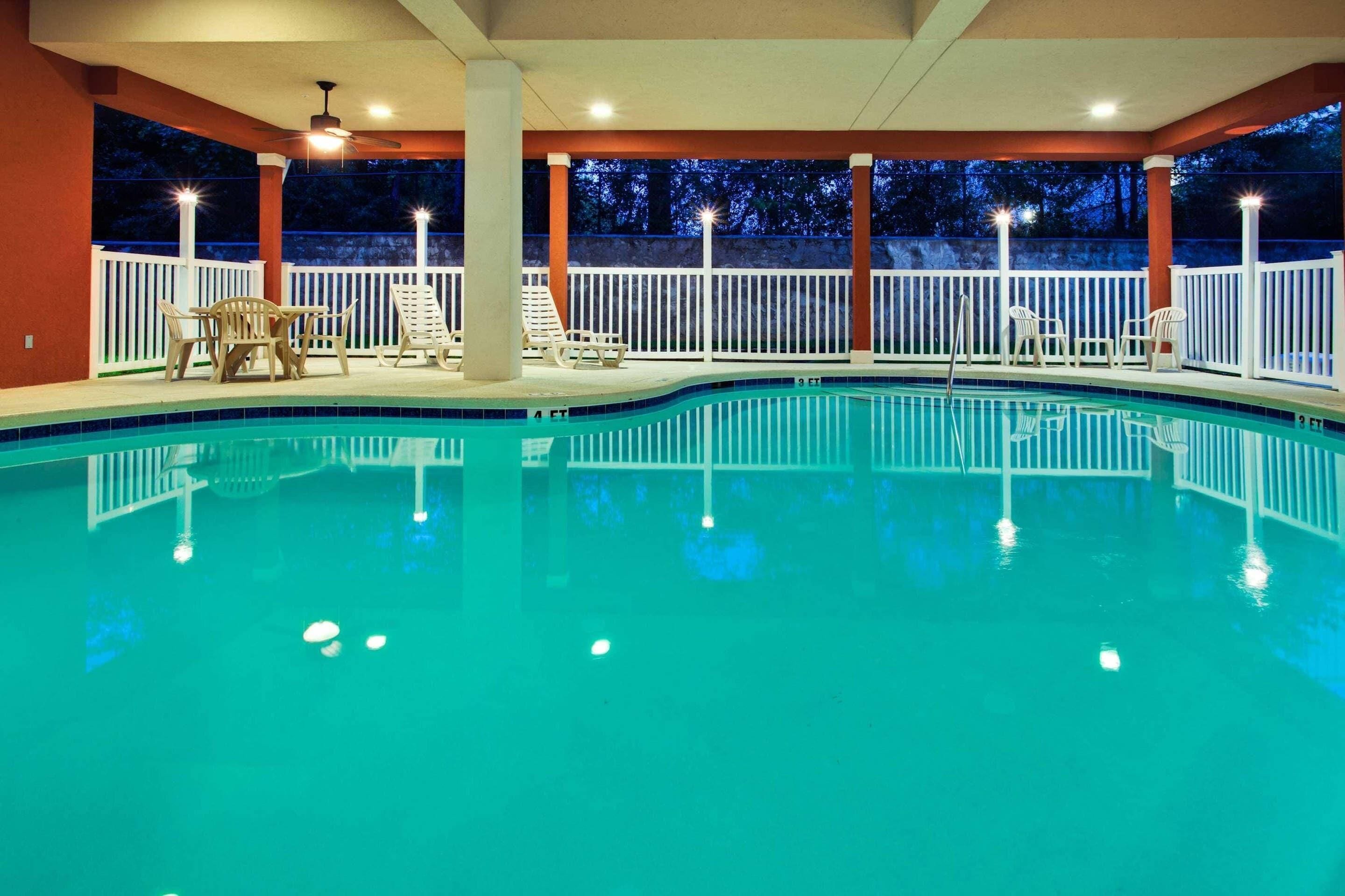 An indoor swimming pool with a white deck, lounge chairs, and a ceiling fan.