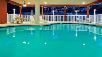 An indoor swimming pool with a white deck, lounge chairs, and a ceiling fan.