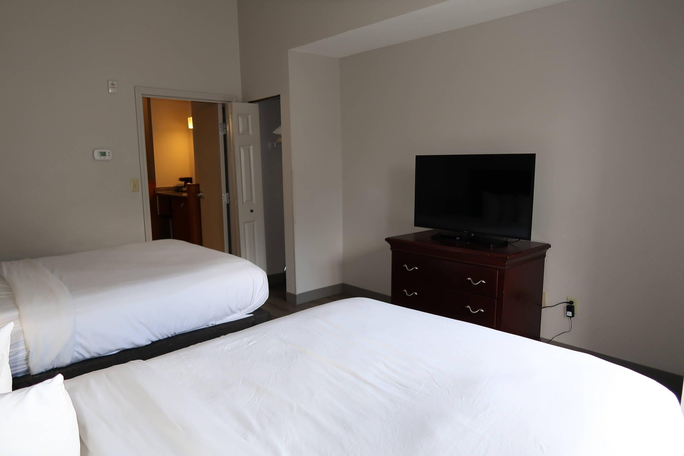 Two single beds with white linens, a television on a wooden dresser, and a door leading to a bathroom.