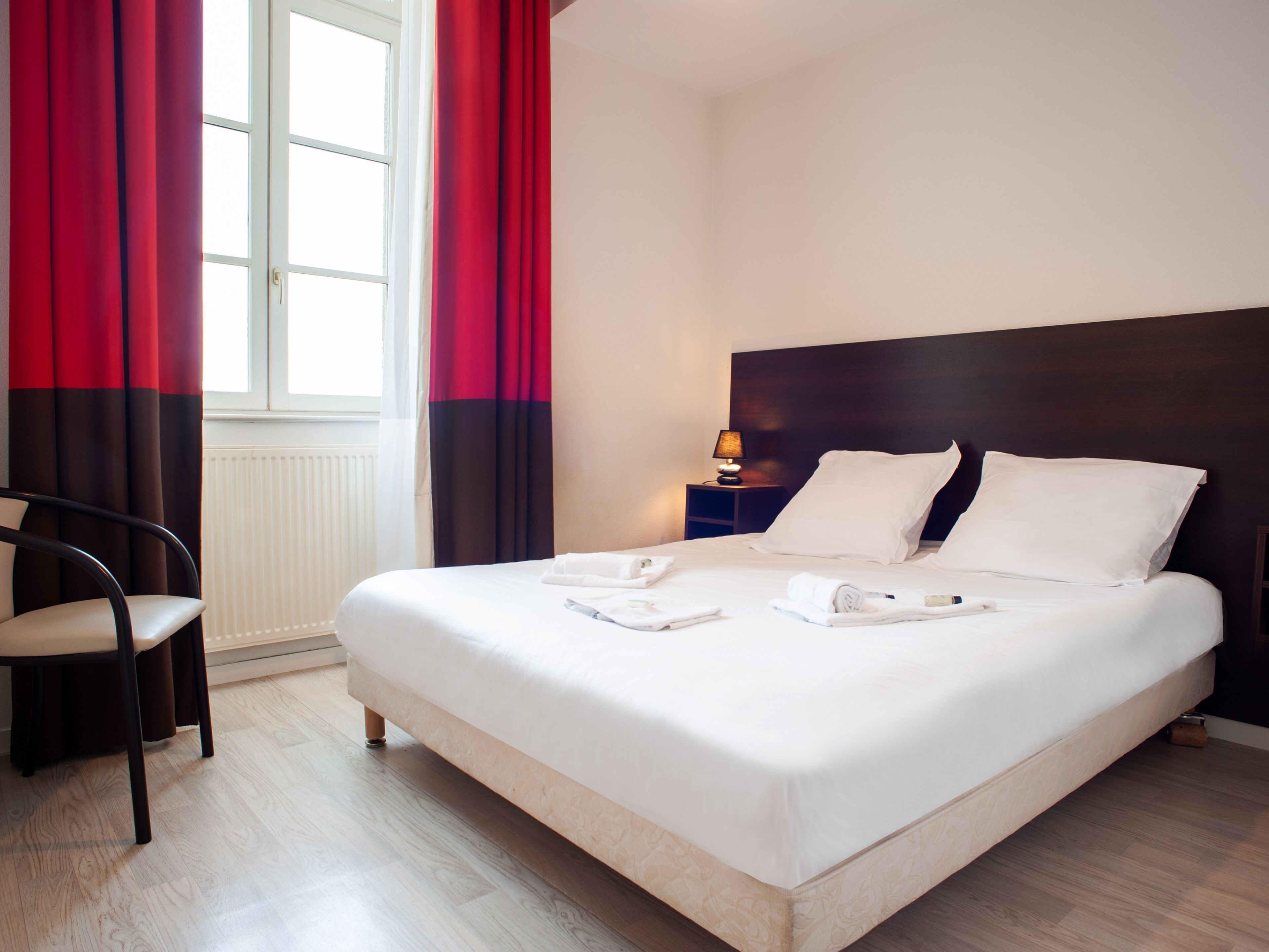 A neatly made bed with white linens, a wooden nightstand with a lamp, and a chair in a room with wooden flooring and a window with red curtains.