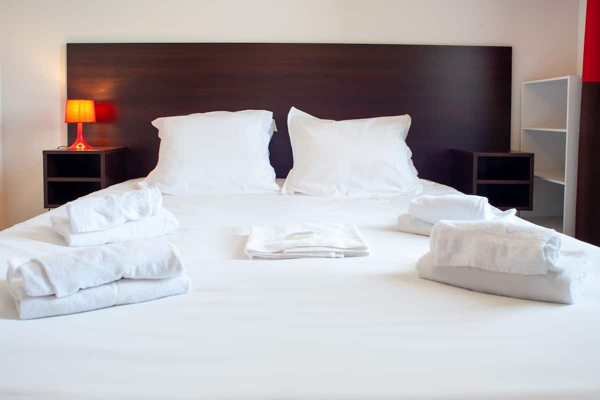 Een netjes opgemaakt bed met witte lakens en kussens, een opgevouwen handdoek in het midden, en een bedlampje aan de linkerkant.