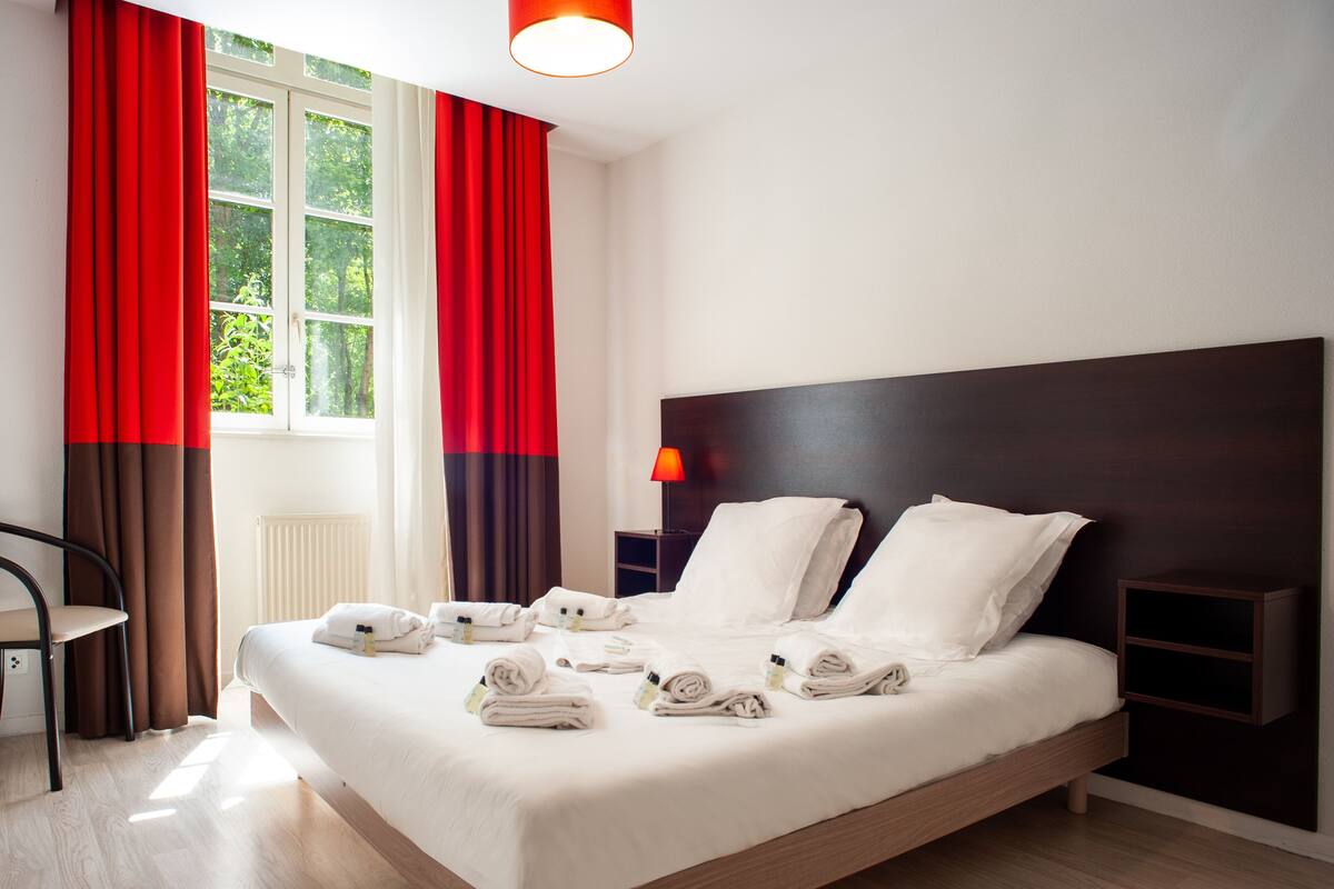 Een keurig opgemaakt bed met witte lakens en handdoeken, een nachtkastje met een lamp, en uitzicht op groen door een raam met rode gordijnen.